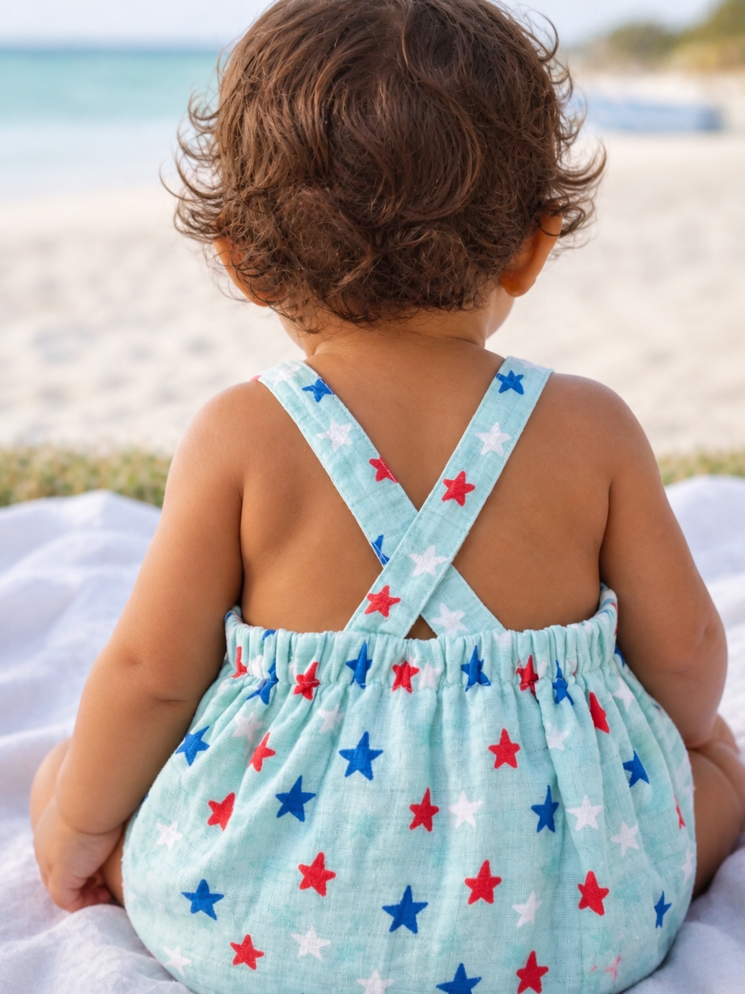 Baby sitting on a blanket, wearing a blue outfit with red and blue stars, facing the beach and ocean.