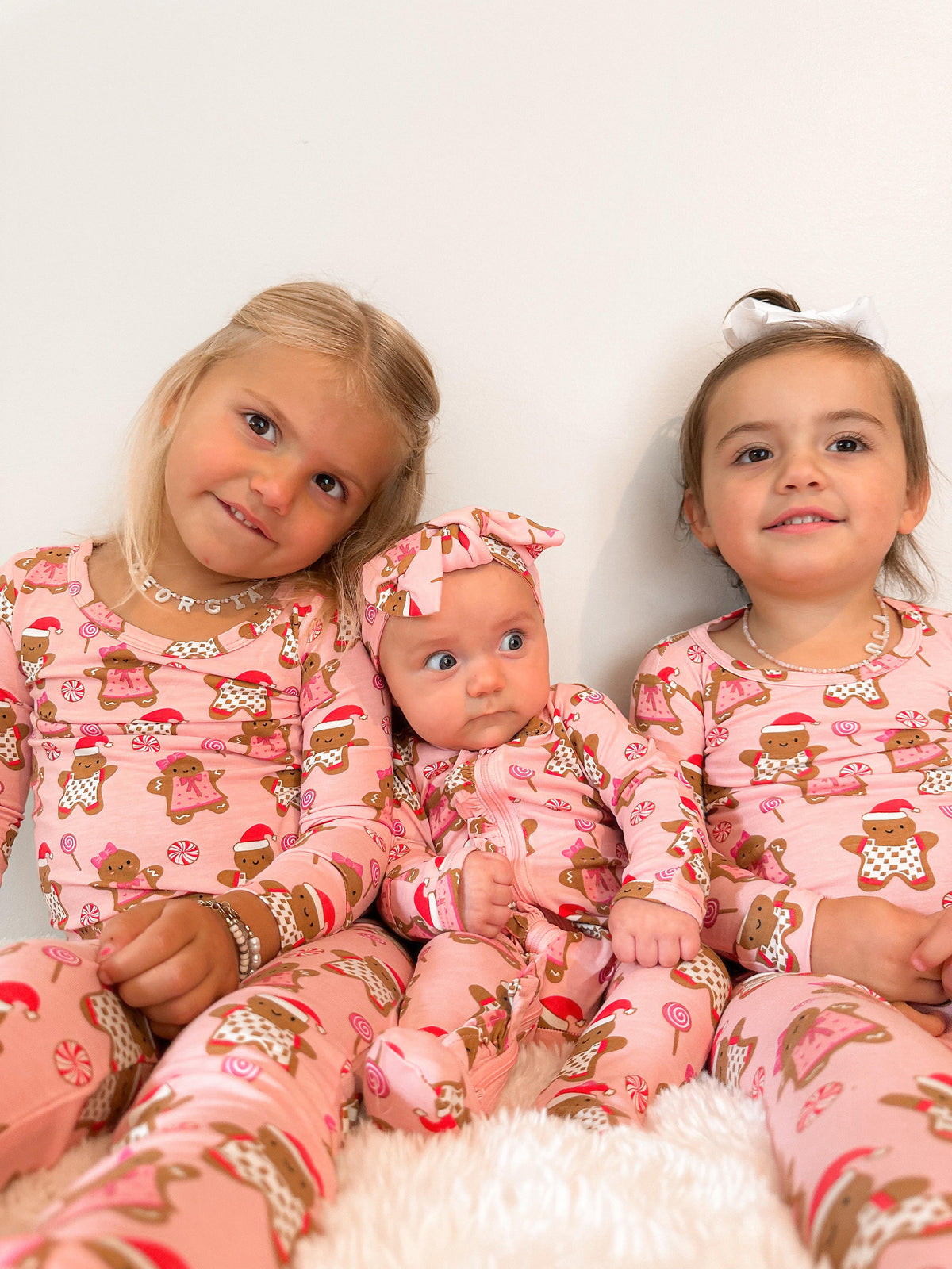 Three young girls in festive pink pajamas with gingerbread motifs, sitting together against a white background.