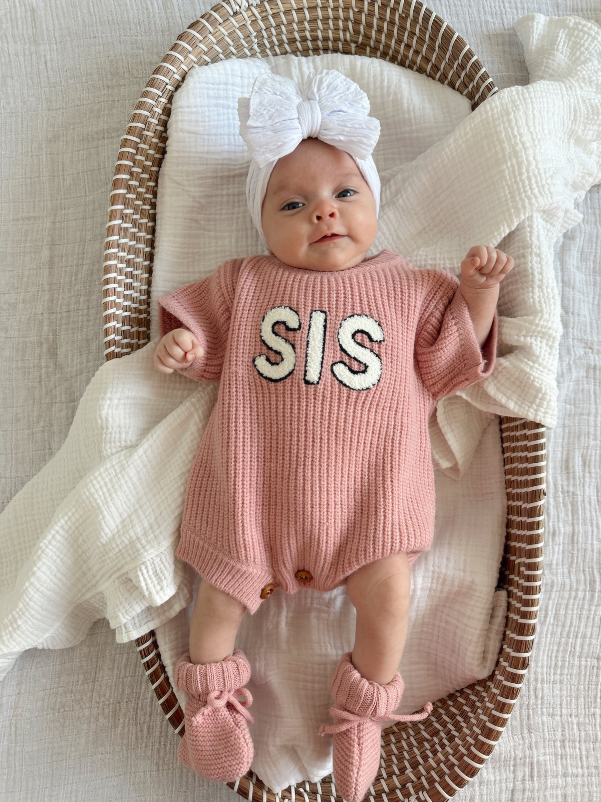 Baby in pink knit romper with "SIS" text, wearing a large white bow and booties, lying in a woven basket.