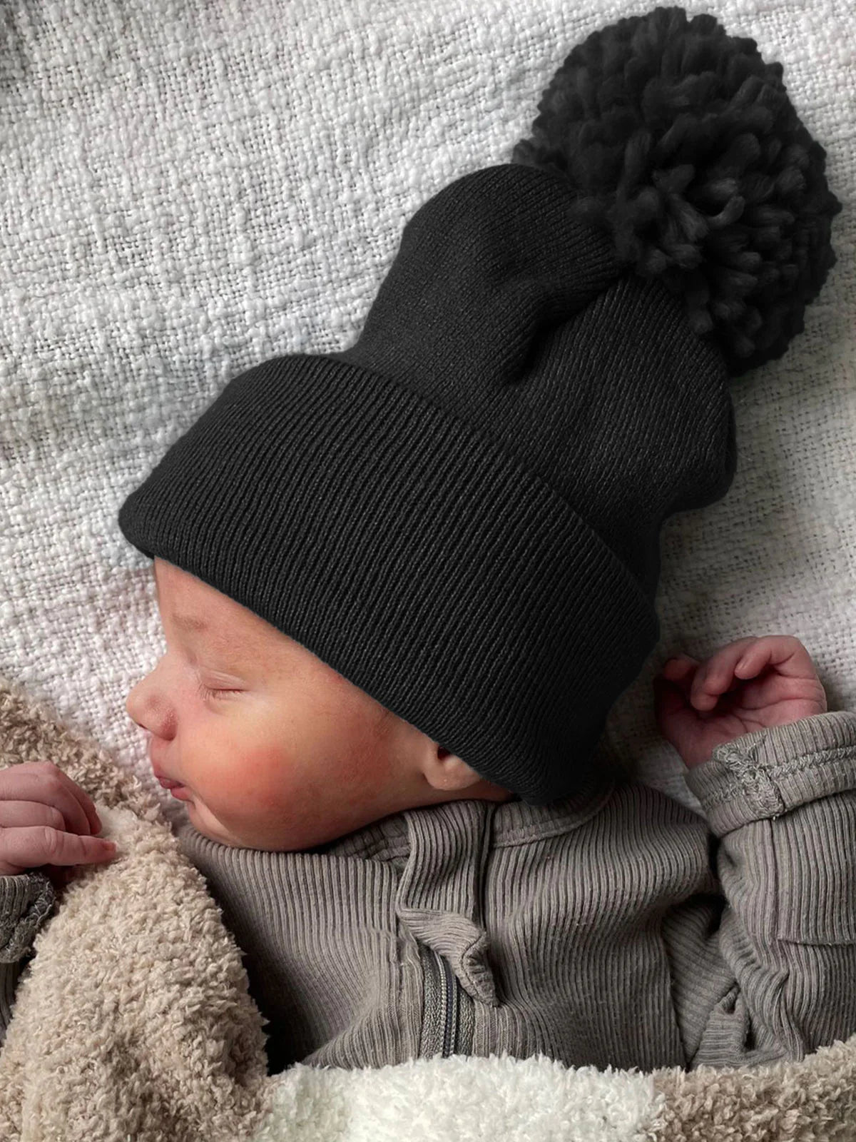 A peaceful newborn in a dark beanie, wrapped in a soft blanket, sleeping on a textured surface.