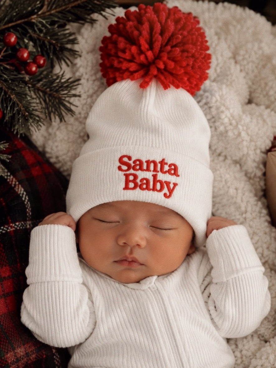 Baby wearing a white "Santa Baby" hat with a red pom-pom, sleeping on a cozy blanket surrounded by festive decor.