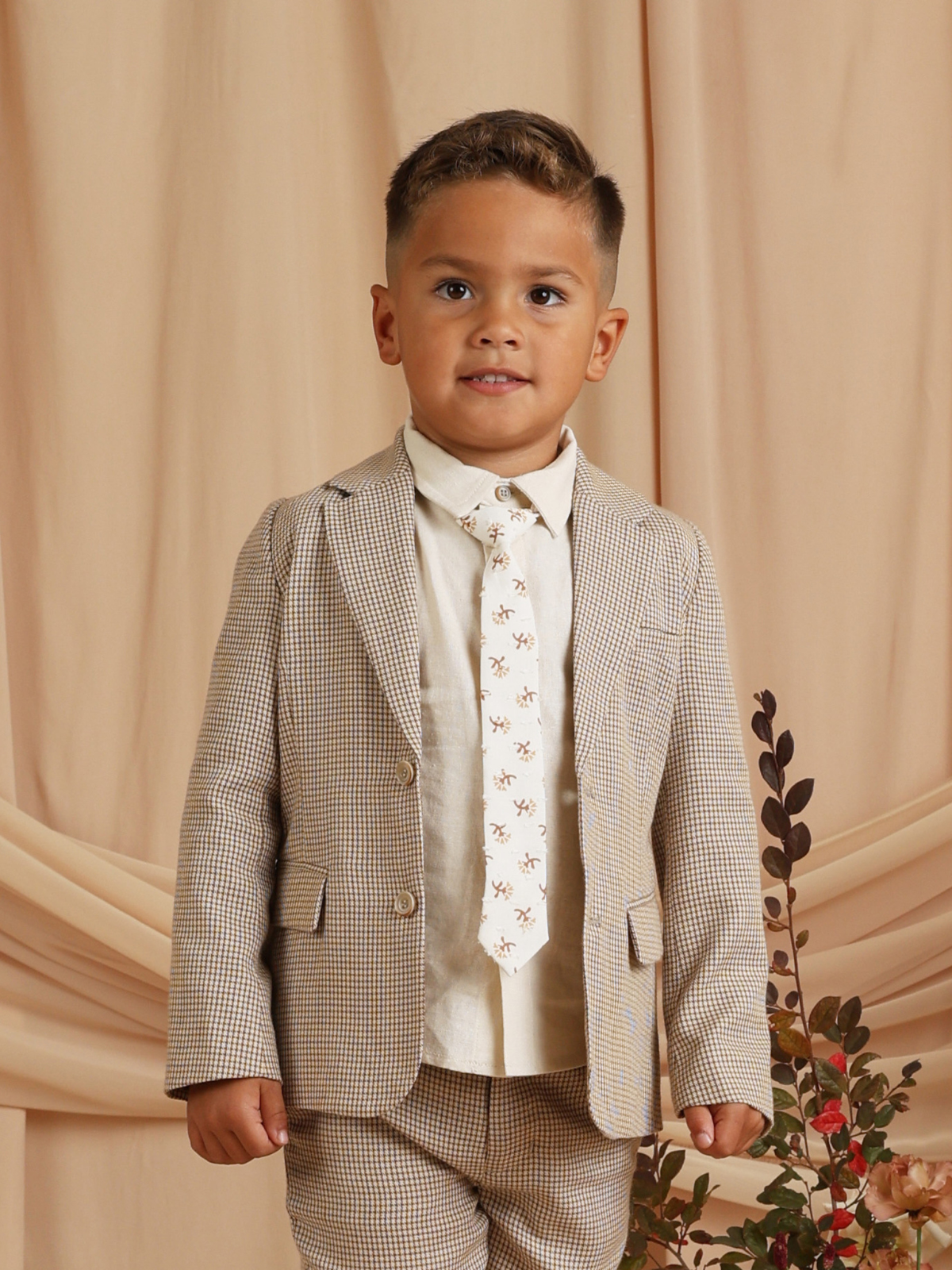 Smiling boy in a checked suit with a patterned tie, standing against a soft, neutral background.