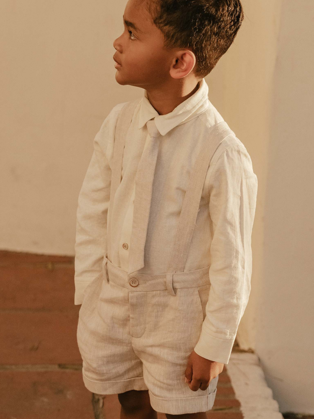 Young boy in cream outfit with suspenders, looking thoughtfully to the side against a soft background.