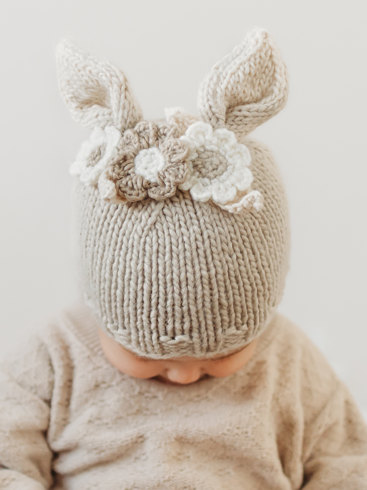 Baby wearing a knitted beige hat with bunny ears and floral embellishments, looking down.