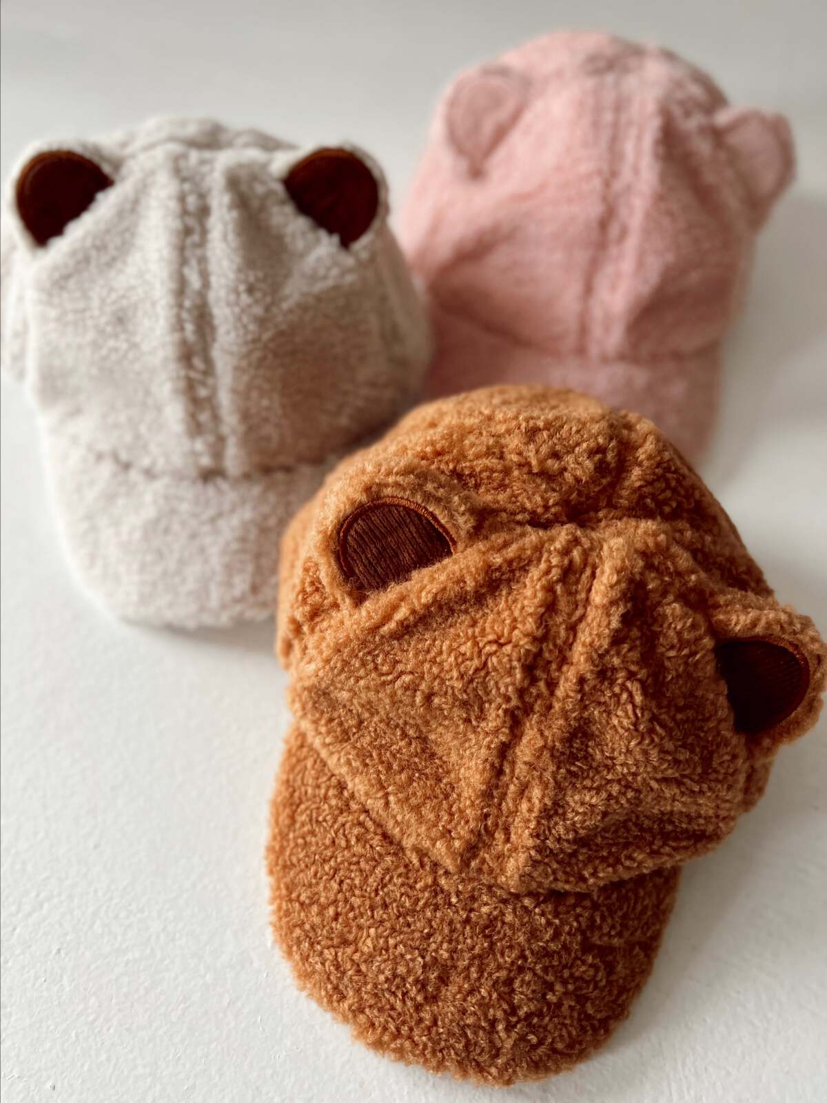 Three fuzzy bear-ear hats in beige, pink, and brown stacked on a white surface.