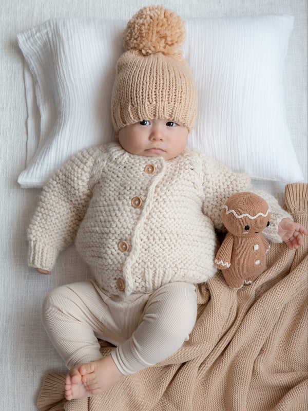 Baby in cozy knitwear and pom-pom hat sits on blanket, with plush toy beside, against a soft backdrop.