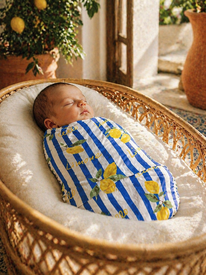 Sleeping baby swaddled in a striped blanket with lemon patterns, nestled in a cozy bassinet indoors.