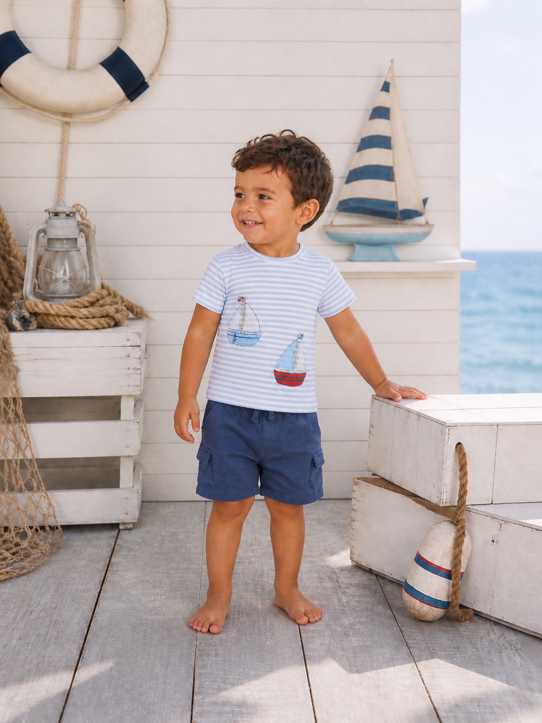 Smiling child in a striped shirt with sailboat designs, standing on a seaside deck with nautical decor.