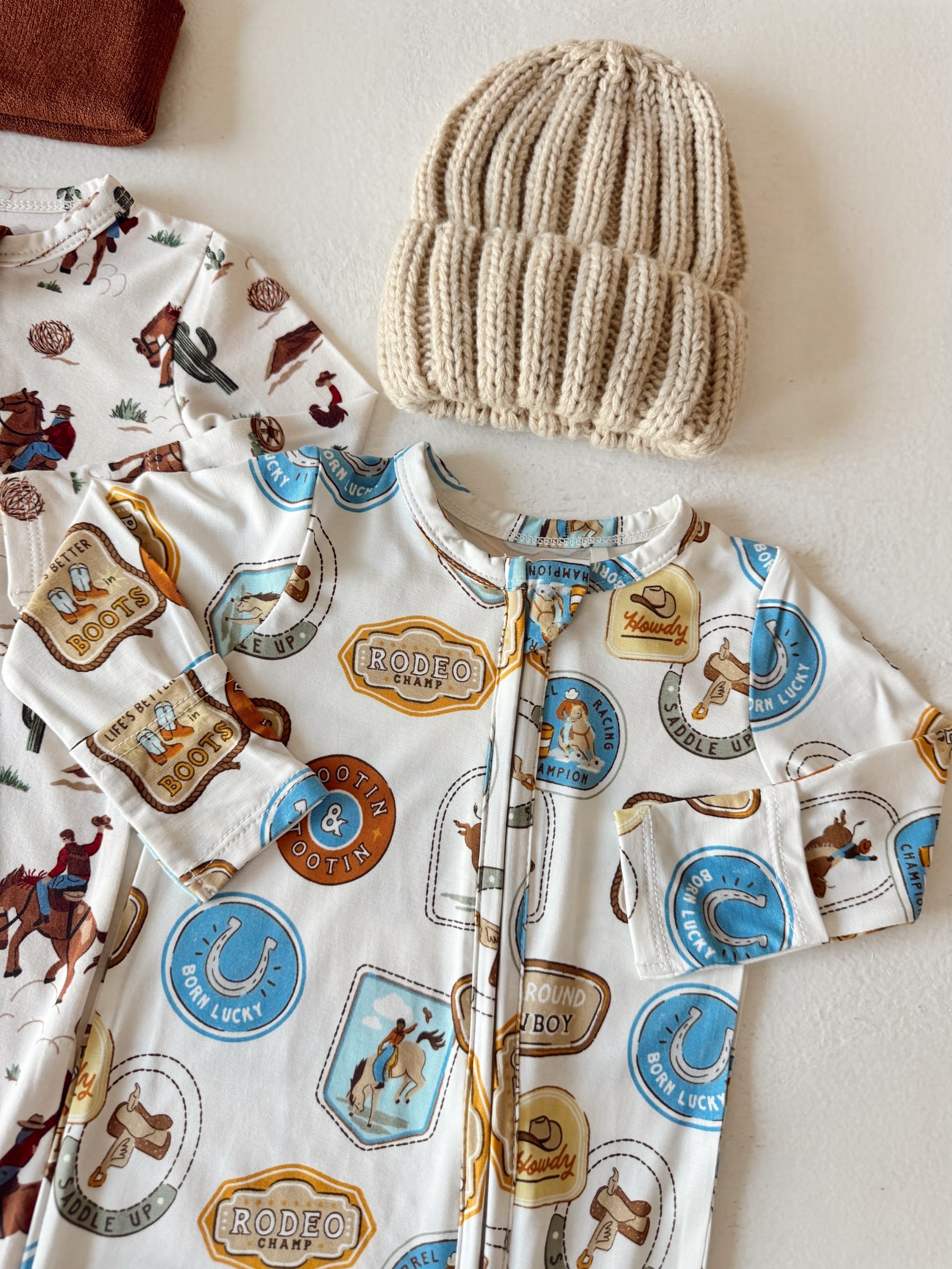 Baby onesie with cowboy print next to a beige knitted hat, resting on a white background.