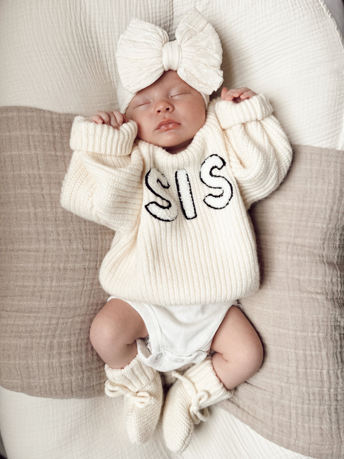 Baby wearing a cream sweater with "SIS" on it, sleeping on a cozy blanket with a large bow headband.
