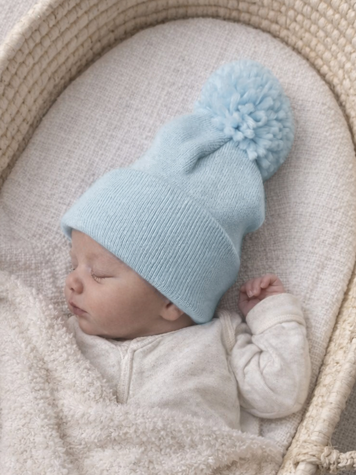 Baby sleeping in a cozy basket, wearing a light blue knitted hat with a pom-pom. Soft, neutral blanket surrounds them.