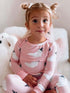 Young girl in pink ghost-patterned pajamas, smiling with playful hairstyle, sitting on soft pillows.