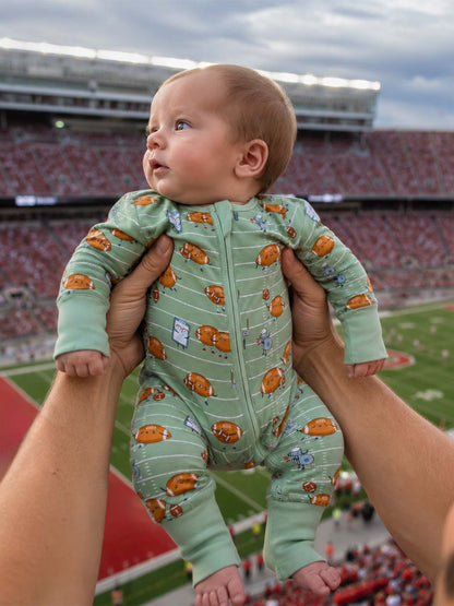 Baby in a green football-themed onesie held up at a stadium with a cheering crowd in the background.