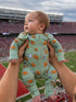 Baby in a green football-themed onesie held up at a stadium with a cheering crowd in the background.