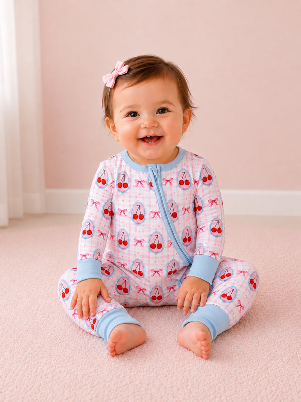 Smiling baby wearing cherry-patterned pajamas, sitting on a soft pink carpet, with a bow in their hair.