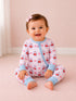 Smiling baby wearing cherry-patterned pajamas, sitting on a soft pink carpet, with a bow in their hair.