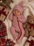 Newborn sleeping in striped red and white outfit, surrounded by gifts and a teddy bear on soft, cozy blanket.