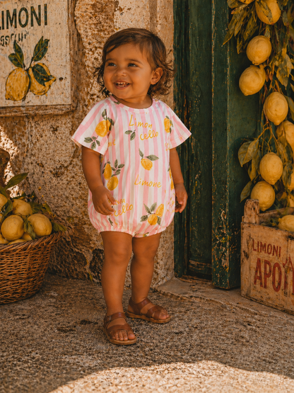 Smiling child in lemon-print outfit stands by a rustic wall adorned with lemons and a basket of fruit.