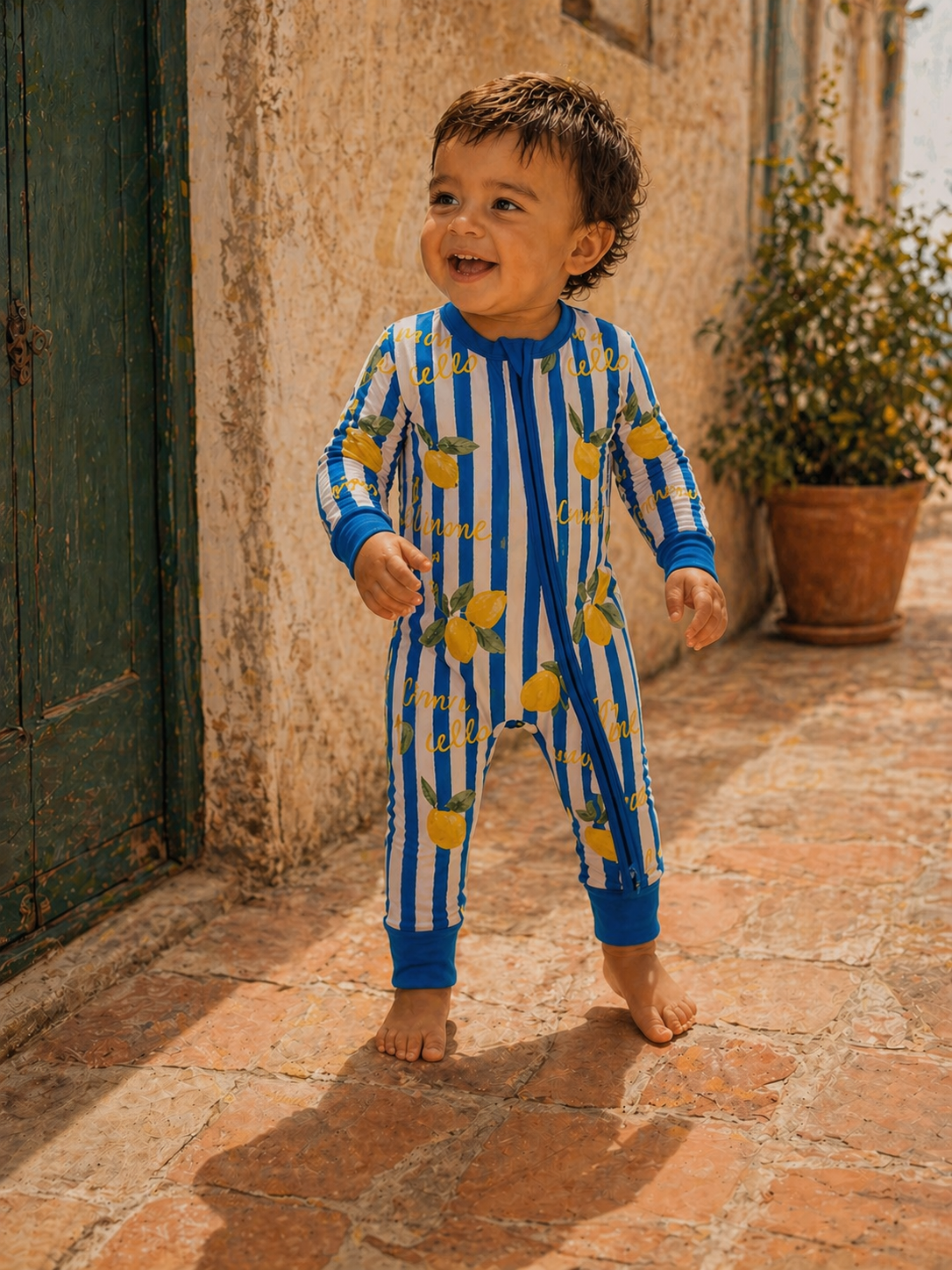 Smiling toddler in lemon-patterned pajamas, standing on a brick pathway near a green door.