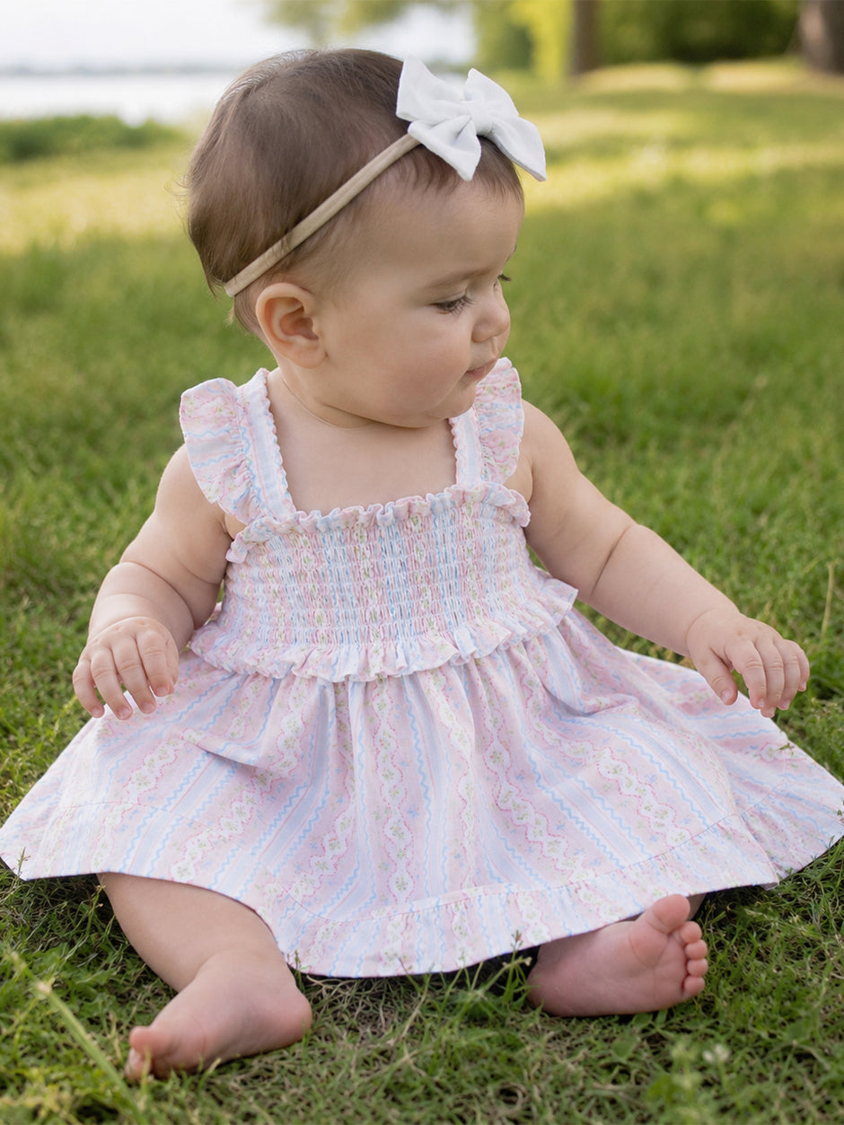 Baby girl sitting on grass, wearing a pastel dress and a headband with a bow, looking to the side.