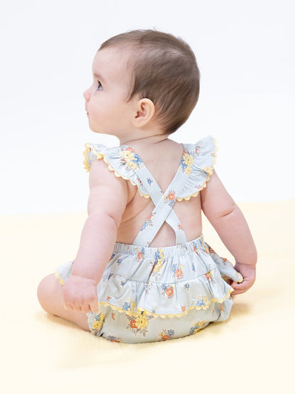 Baby in a floral dress with ruffles, sitting on a yellow background, looking back over their shoulder.