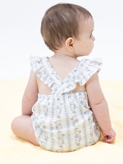 Baby in a floral romper sits on a soft yellow blanket, turned away, showcasing the outfit's back design.