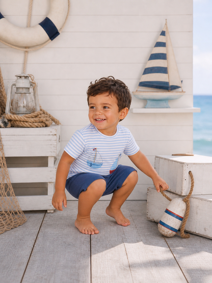 Smiling boy in striped shirt squats near nautical decor on a sunny beach.