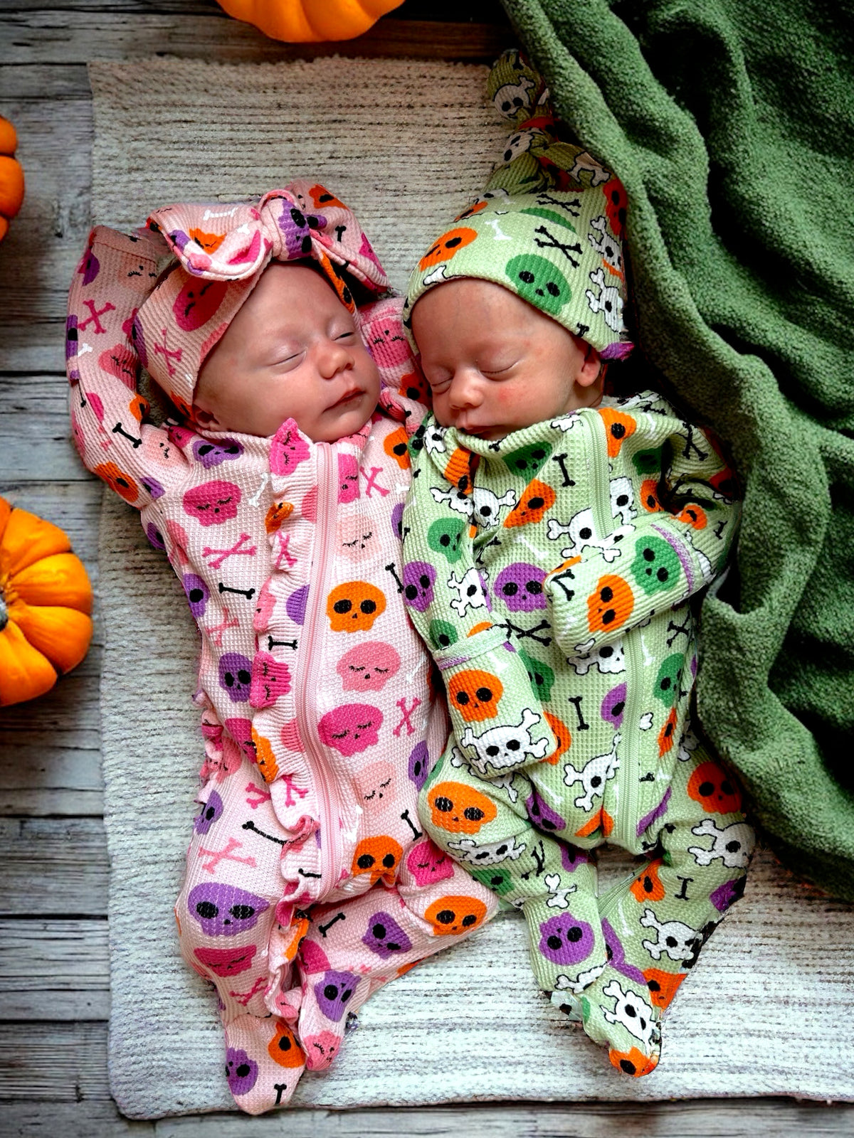 Two newborns dressed in Halloween-themed onesies, sleeping on a textured blanket surrounded by pumpkins.