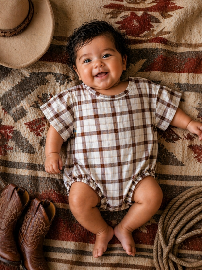 Smiling baby in a plaid outfit, surrounded by cowboy boots and a hat on a textured blanket.