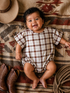 Smiling baby in a plaid outfit, surrounded by cowboy boots and a hat on a textured blanket.