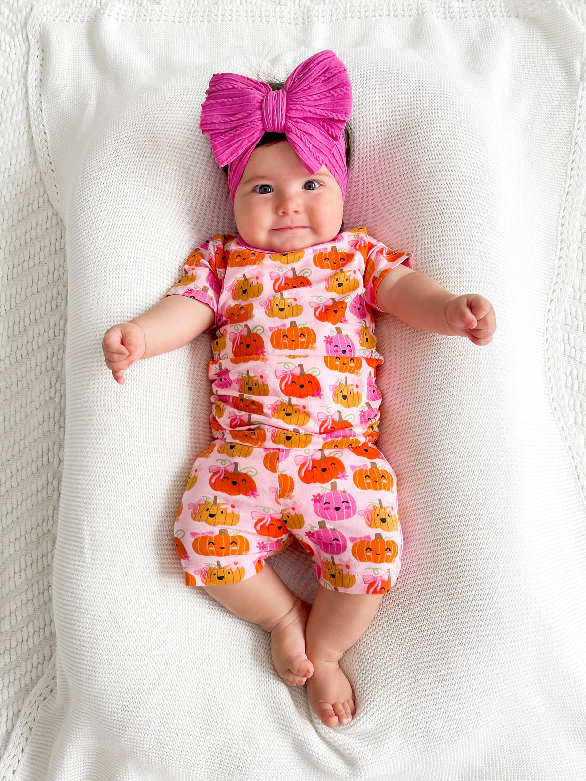 Infant with a large pink bow, wearing a pumpkin-patterned outfit, lying on a white blanket.