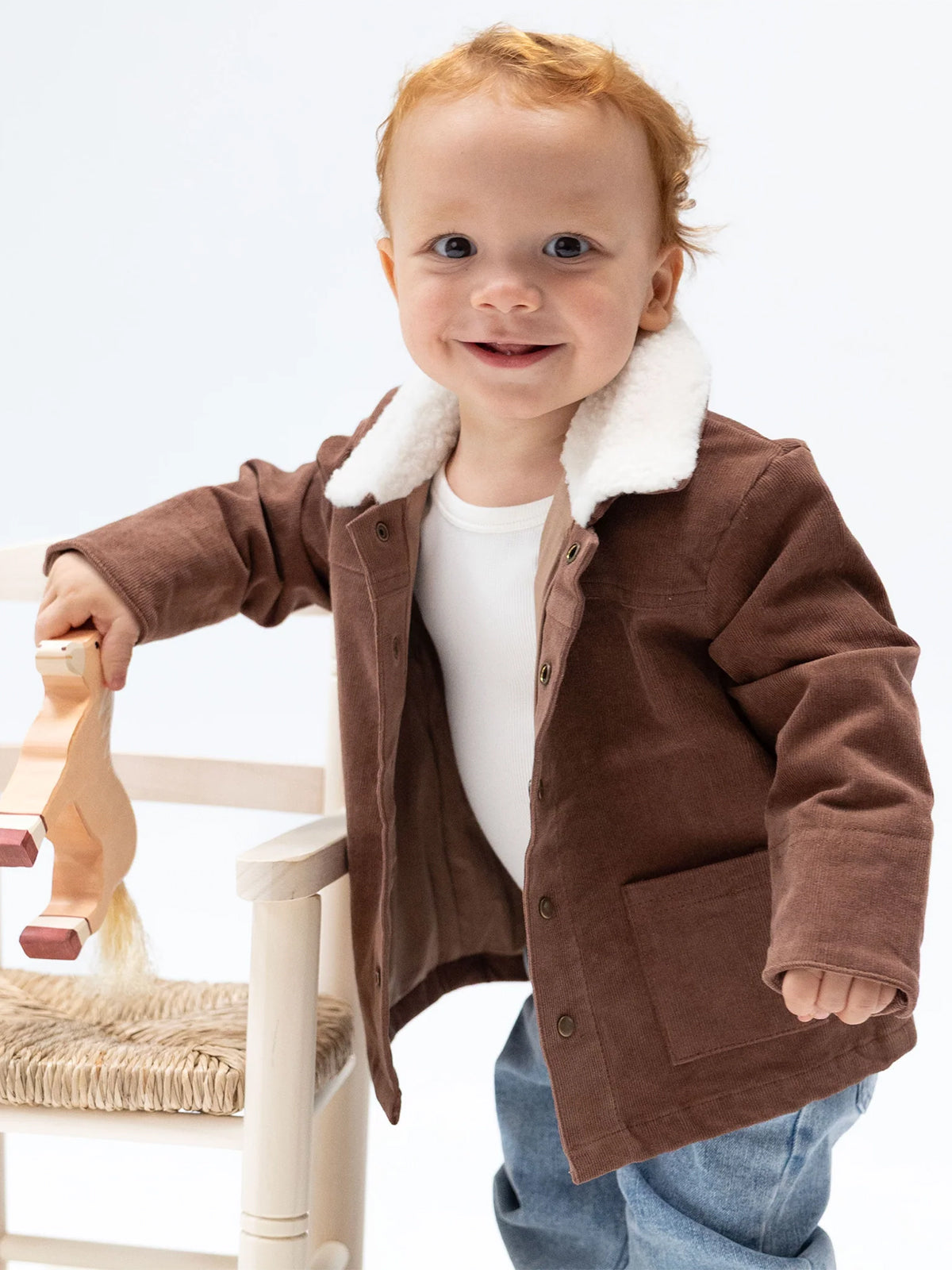 Smiling toddler in a brown jacket holding a toy horse, standing next to a chair against a white background.