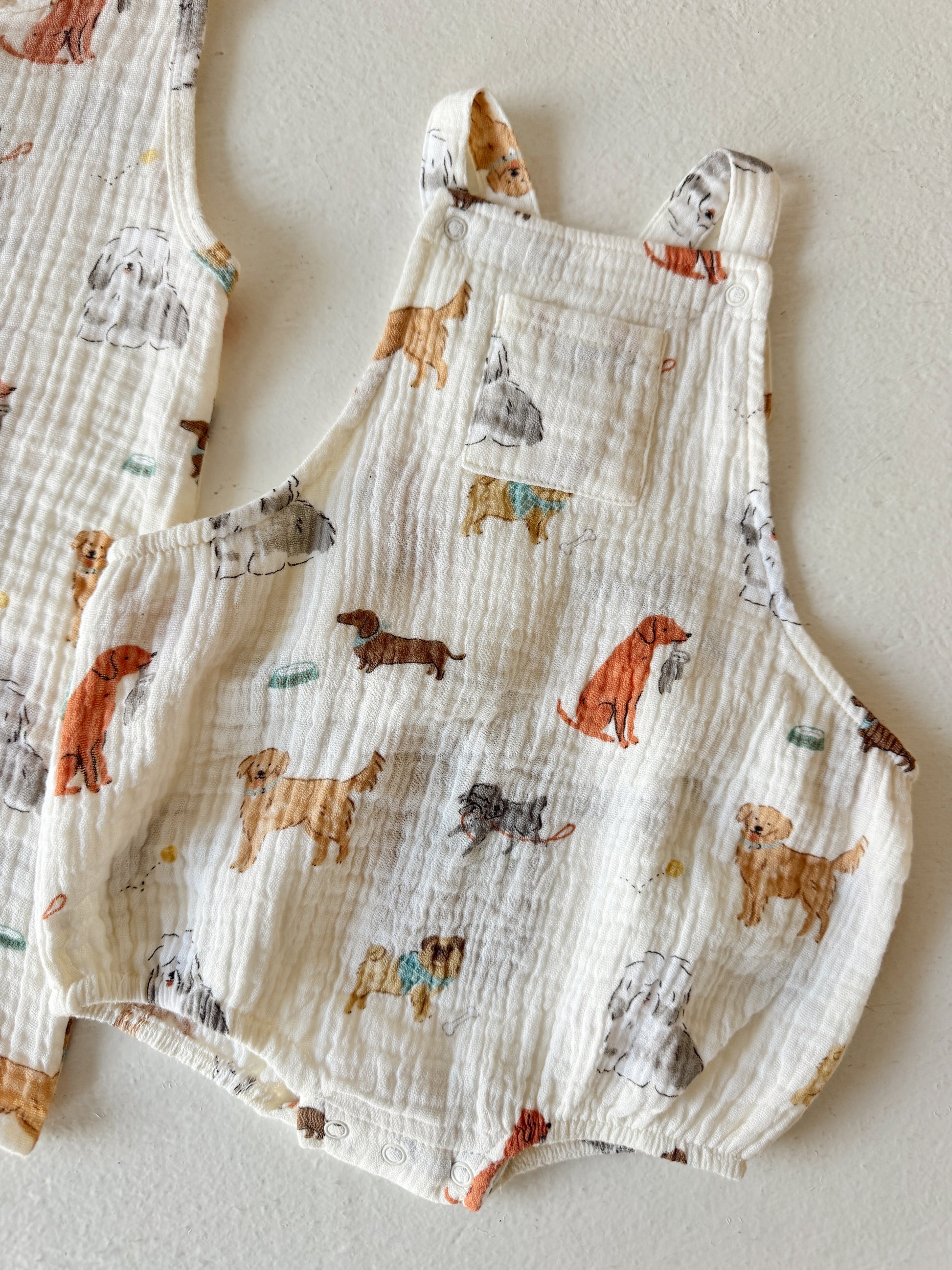 White fabric with playful dog patterns, featuring various breeds and a pocket detail on overalls.