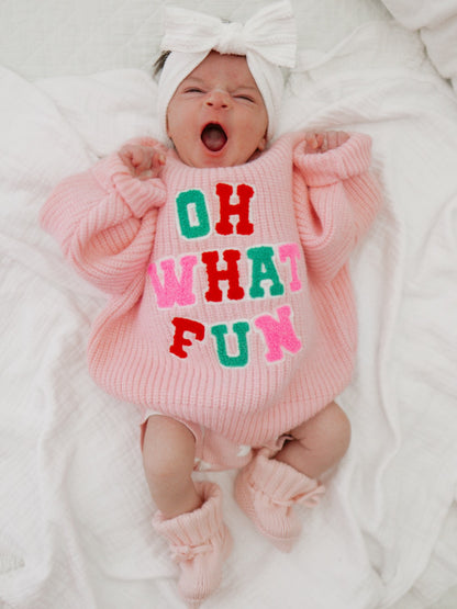 Baby yawning in a pink sweater with "OH WHAT FUN" text, wearing a headband and cozy booties, on a white blanket.