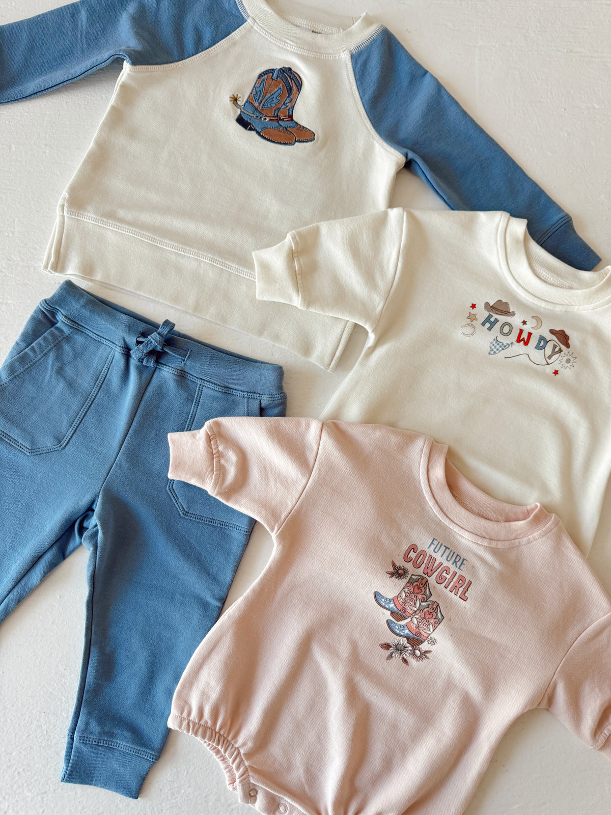 Kids' western-themed apparel: two sweatshirts, a onesie, and pants featuring cowboy motifs and playful designs.