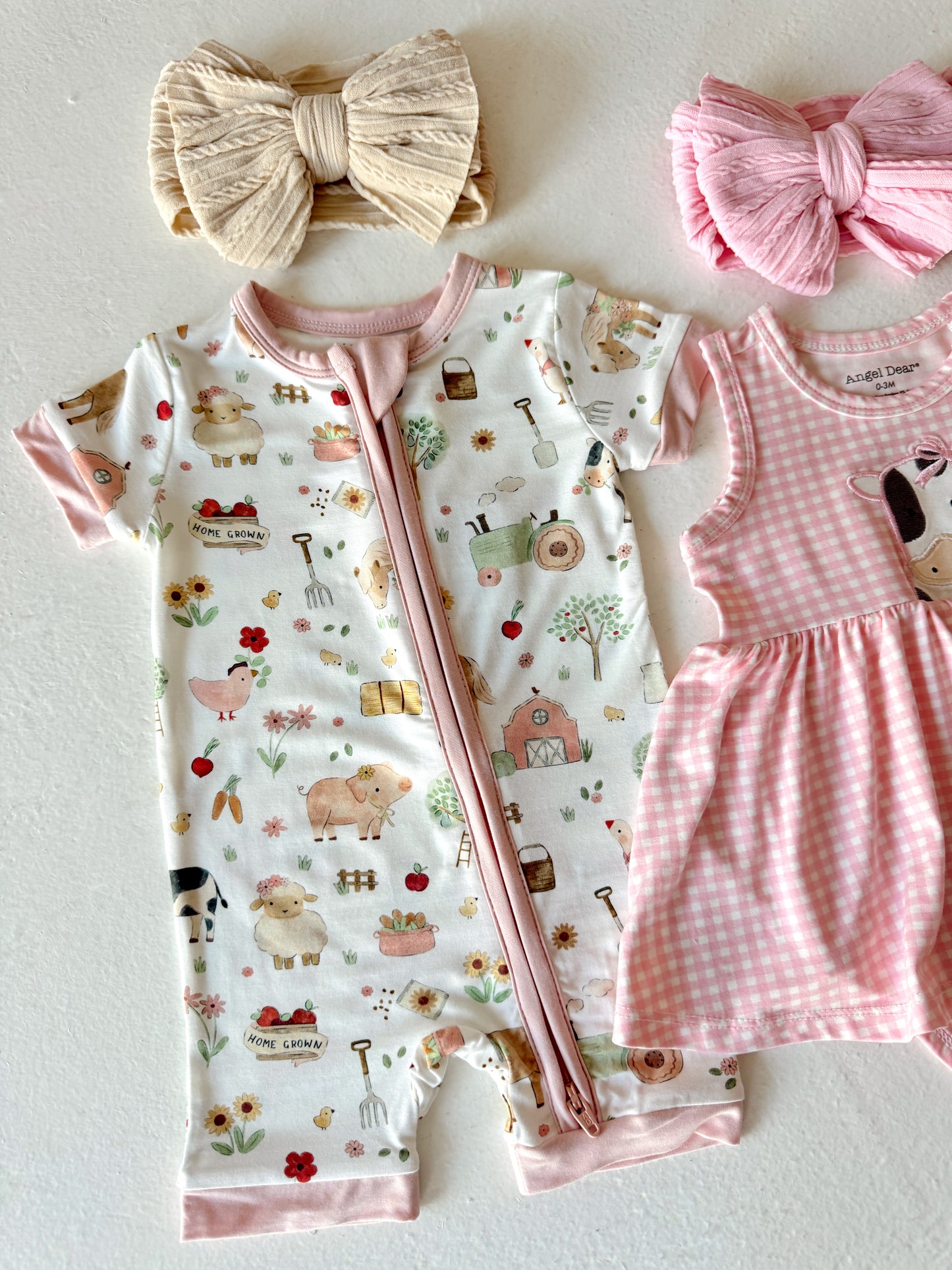 Toddler clothing: a farm-themed onesie, pink gingham dress, and two hair bows in cream and pink.