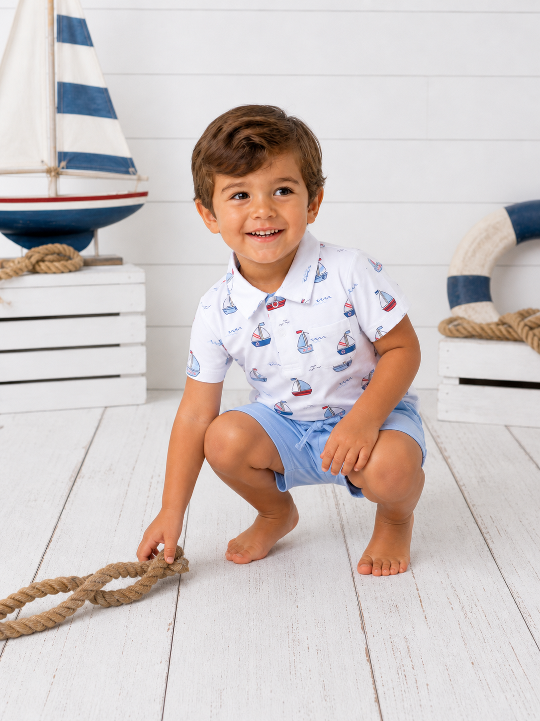 Smiling child in a boat-patterned shirt squatting on a wooden floor, playing with a rope in a coastal setting.