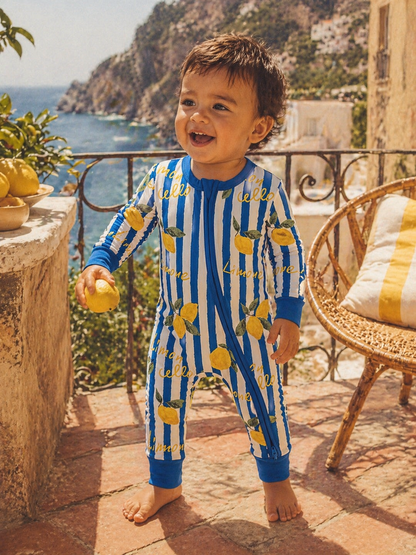 Smiling child in a lemon-patterned onesie, holding a lemon by a coastal terrace with greenery and a view of the sea.