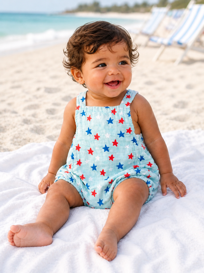 Smiling baby in a star-patterned romper seated on a beach towel, with a sandy beach and ocean in the background.