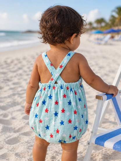 Child in a star-patterned romper stands on the beach, facing the ocean, with beach chairs in the background.