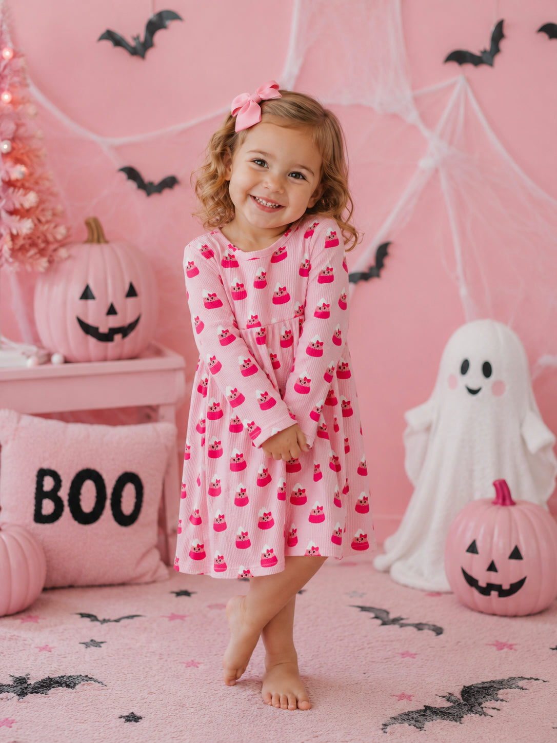 Smiling child in pink cupcake dress stands in a festive Halloween setting with pumpkins and decorations.