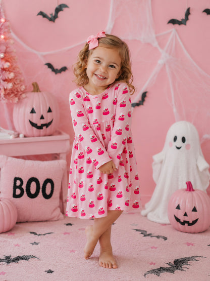 Smiling child in pink cupcake dress stands in a festive Halloween setting with pumpkins and decorations.