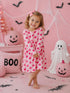 Smiling child in pink cupcake dress stands in a festive Halloween setting with pumpkins and decorations.