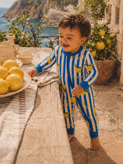 Toddler in striped lemon pajamas smiles beside a table of lemons and a scenic coastal background.