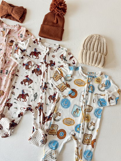 Three baby rompers with cowboy prints, along with two knitted hats in brown and cream, laid on a white surface.