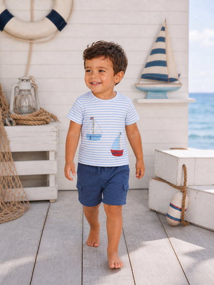 Smiling boy in a striped shirt with boat prints, walking on a wooden deck by the beach.