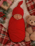 Newborn swaddled in red, sleeping peacefully beside a teddy bear on a cozy plaid blanket and festive decor.