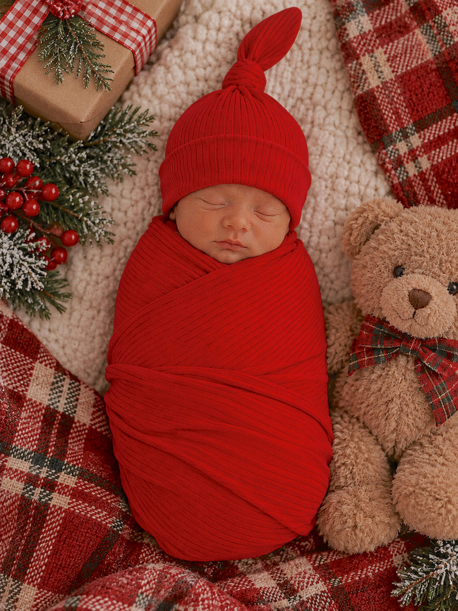 Newborn swaddled in red, sleeping peacefully beside a teddy bear on a cozy plaid blanket and festive decor.