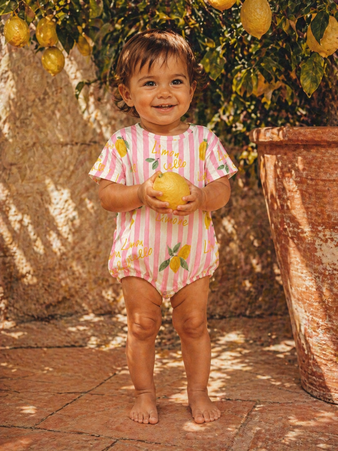 Smiling child in lemon-themed outfit holding a lemon, standing near a lemon tree in a sunny outdoor setting.