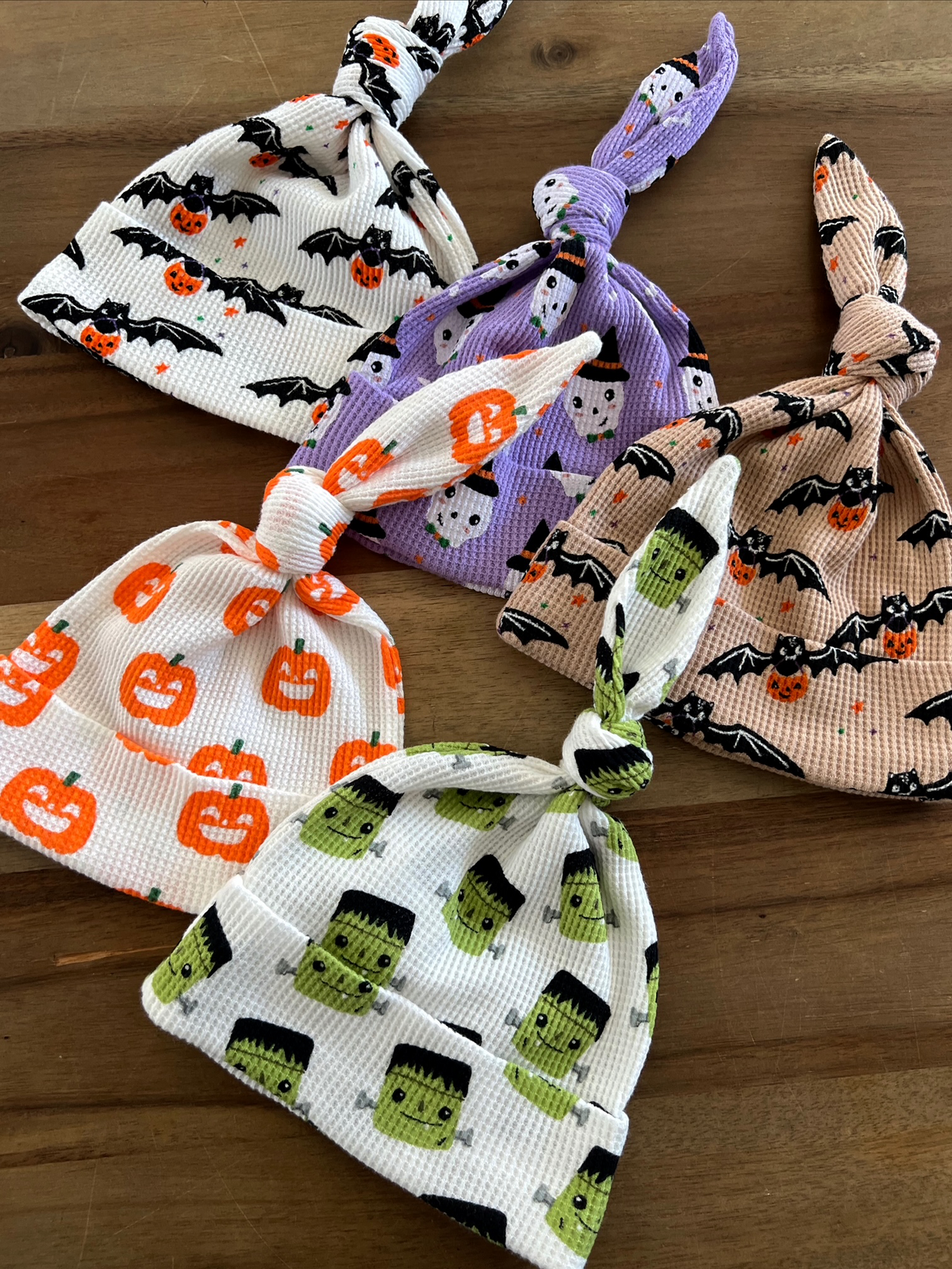 Four Halloween-themed knotted baby hats featuring pumpkins, ghosts, and Frankenstein characters on a wooden surface.